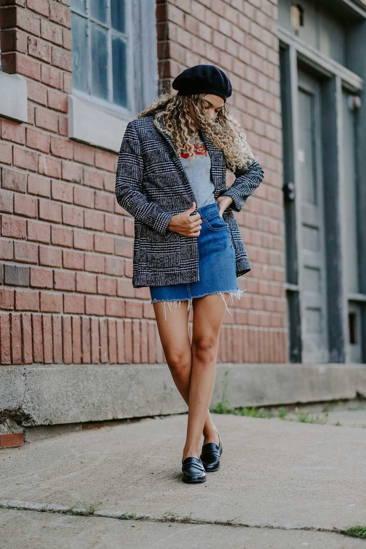 Woman posing in a plaid blazer, denim skirt, and black loafers against a brick wall.