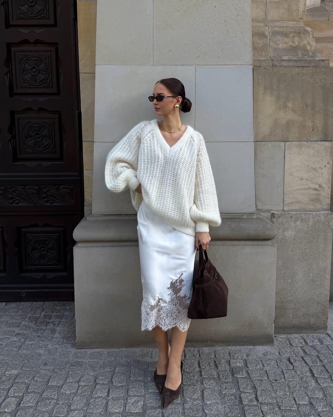 Woman wearing a white cable-knit sweater and a white satin lace skirt standing outdoors.