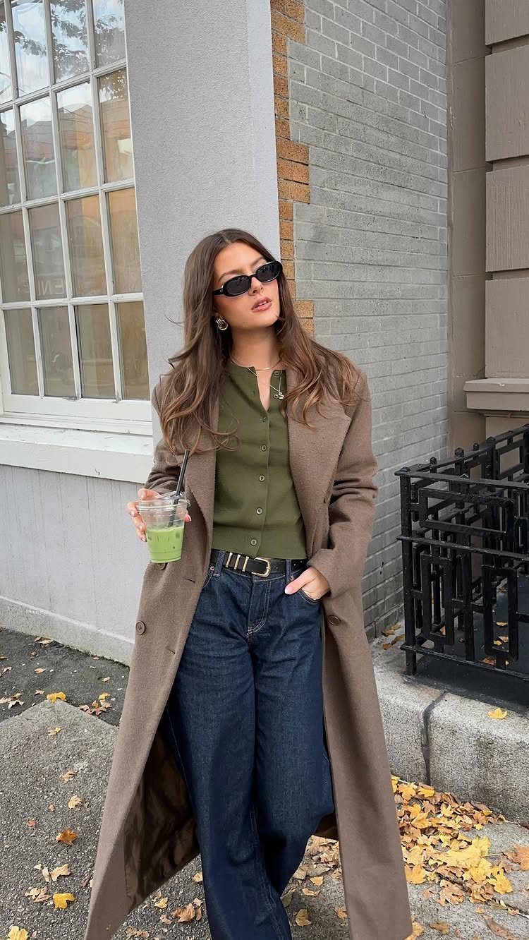 Woman wearing a long brown coat, olive green shirt, and dark blue jeans standing outdoors.