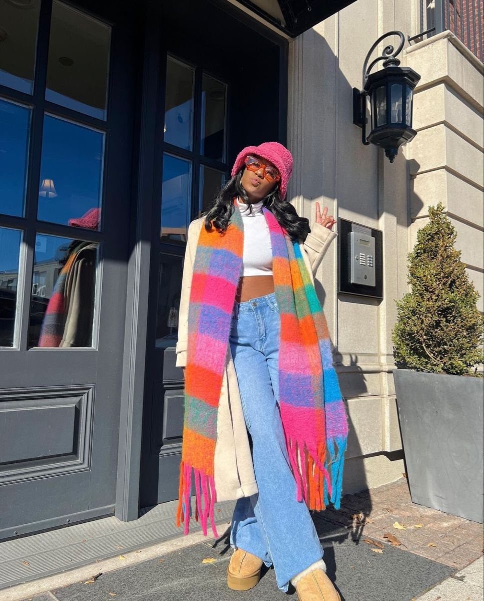 A woman posing in a bright, multi-colored striped scarf, pink beanie, and blue jeans.
