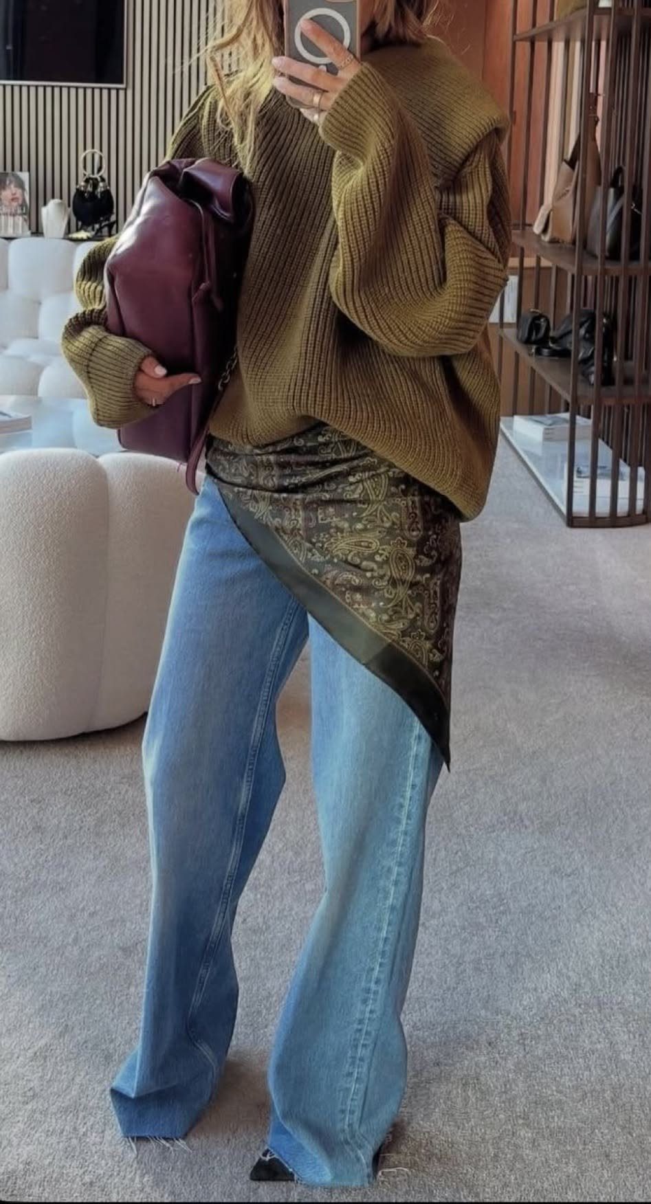 Woman in olive knit sweater, blue jeans, and a patterned waist scarf holding a purple bag.