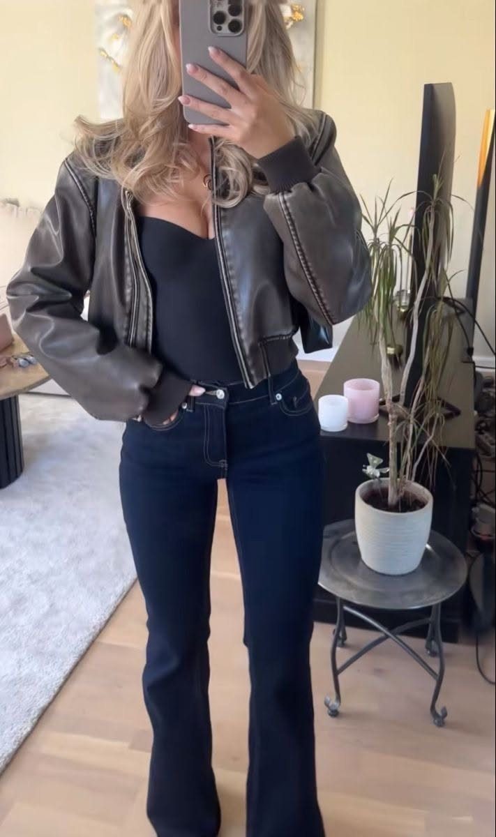 A woman posing in a mirror wearing a brown leather jacket, black bodysuit, and flared jeans.