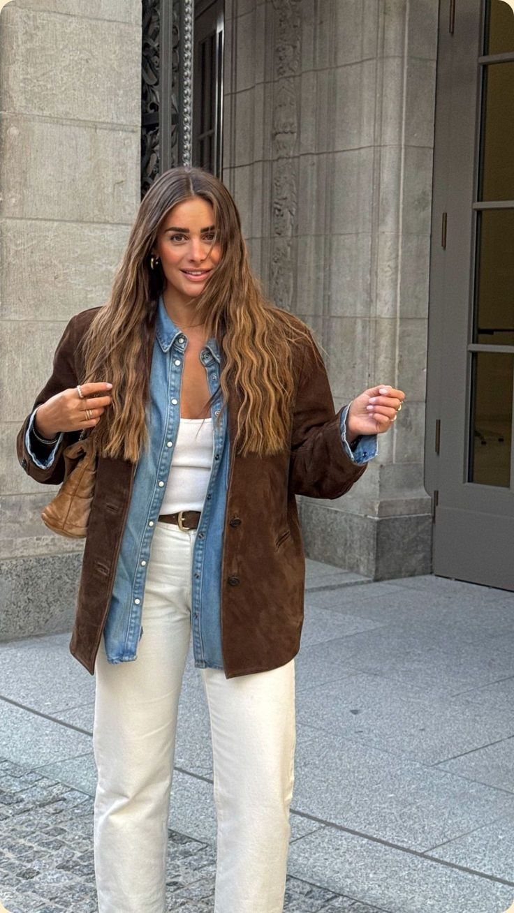 Woman wearing a brown suede jacket, denim shirt, and white trousers standing outdoors.