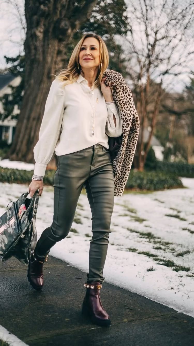 Woman walking in snow wearing white blouse, green leather pants, and purple lug sole boots.