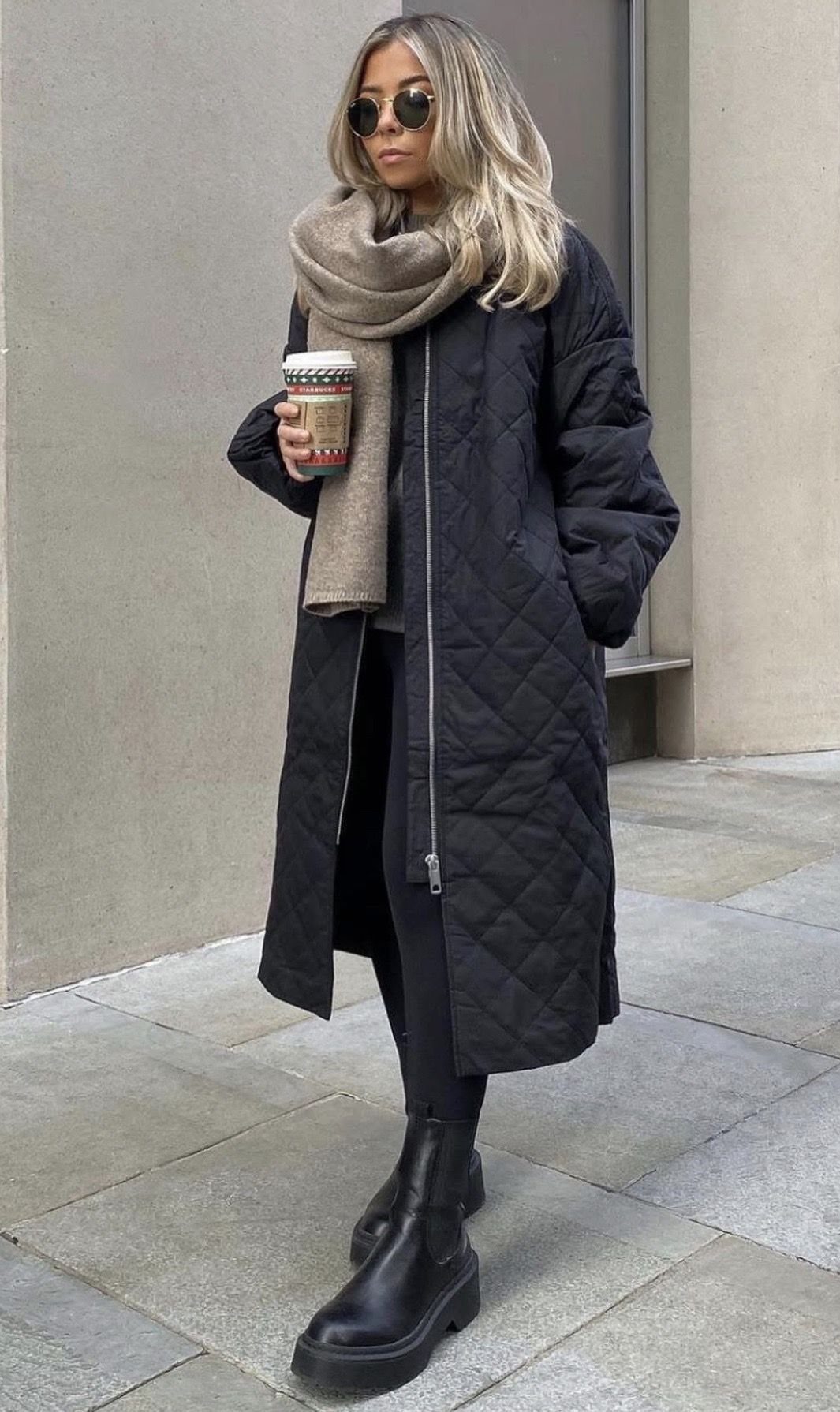 Woman in a long black quilted jacket, beige scarf, and black boots standing outdoors.