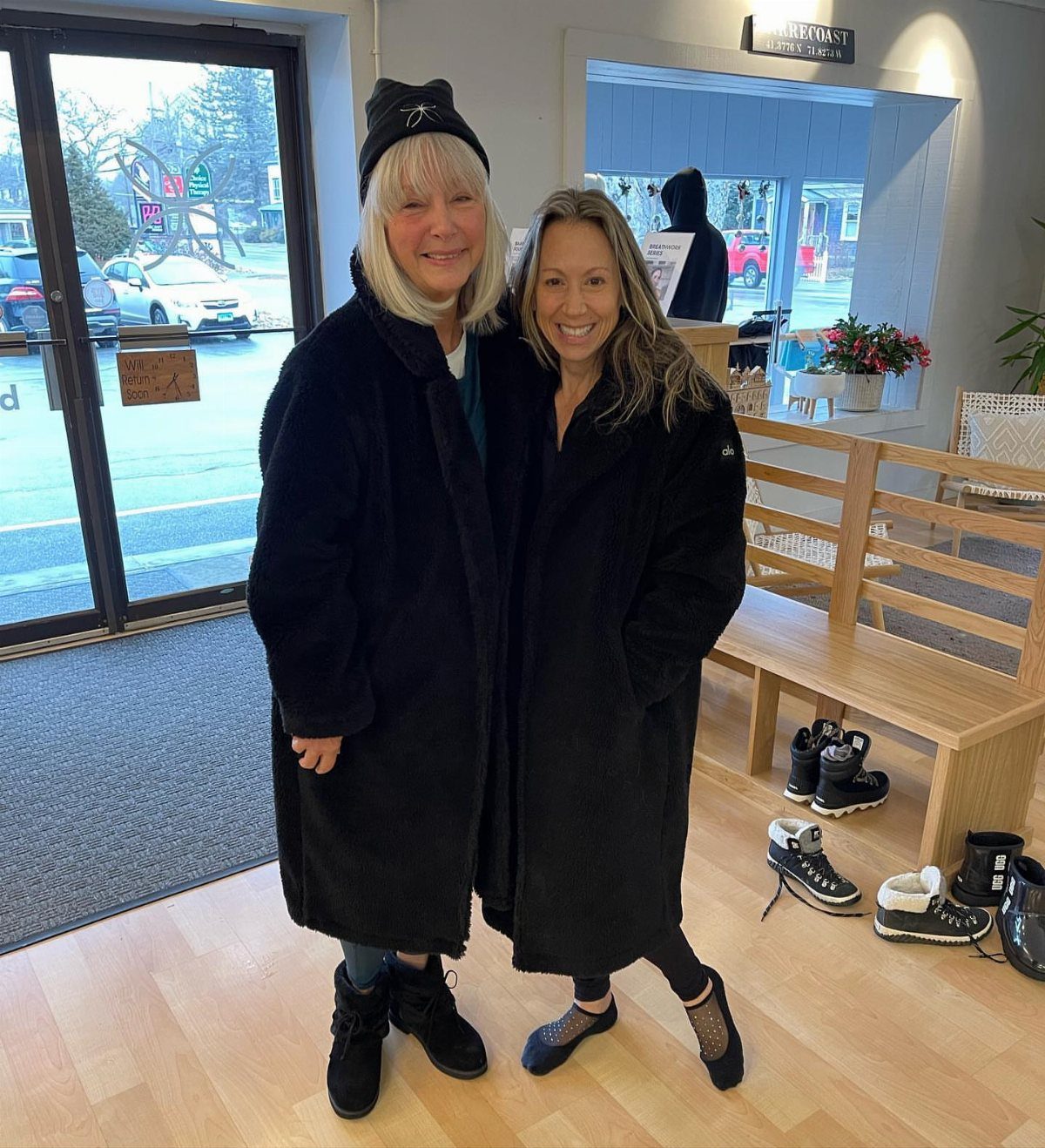 Two women smiling while wearing long black fuzzy sherpa coats in a shop.