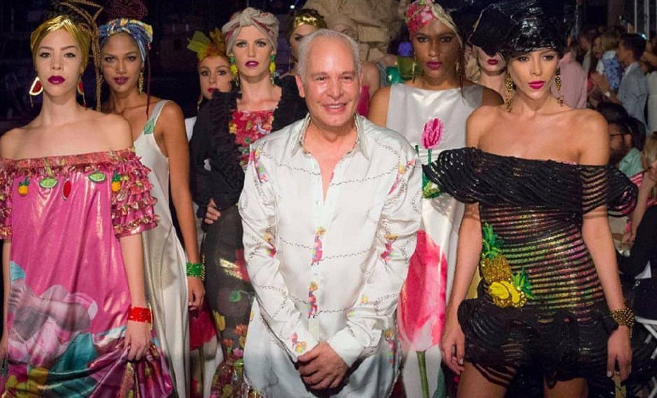 A male designer smiling among models wearing colorful, patterned high-fashion clothing at a runway show.