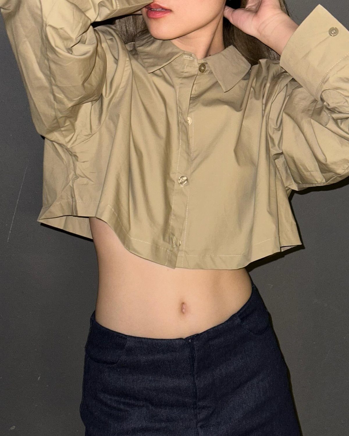 A person wearing a tan cropped button-up shirt and dark blue trousers against a grey wall.