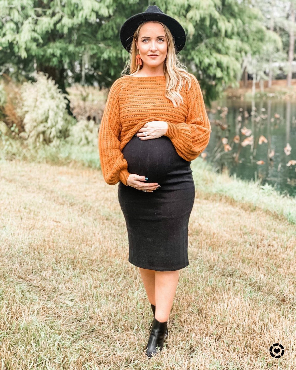 Pregnant woman in an orange cropped sweater and black skirt standing in a grassy field.