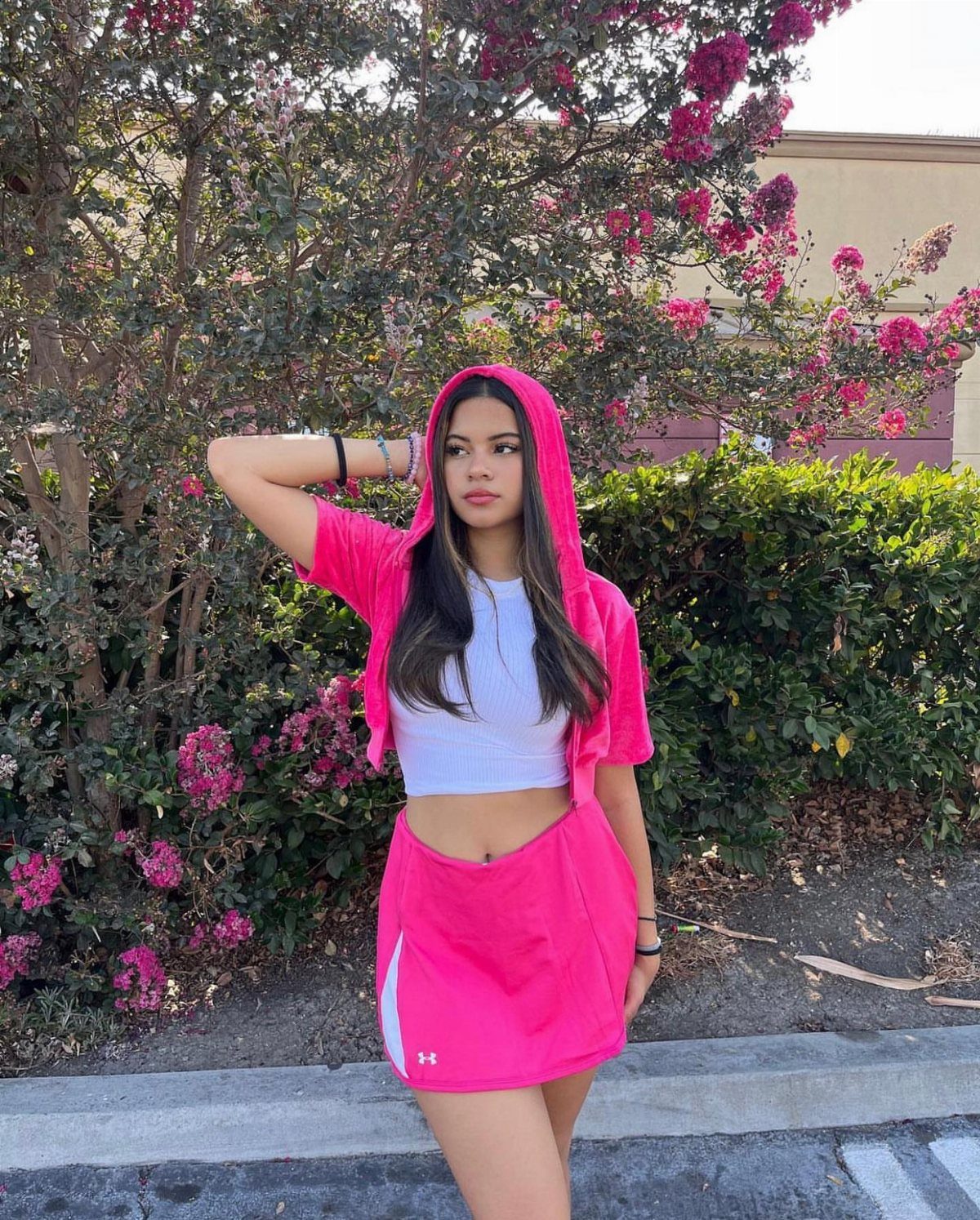 Young woman wearing a white cropped tank top and a pink skirt standing by bushes.