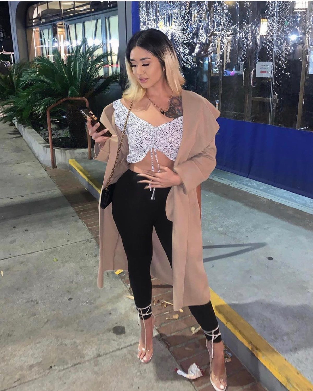 A woman in a sequined crop top, tan coat, and black leggings posing outdoors at night.