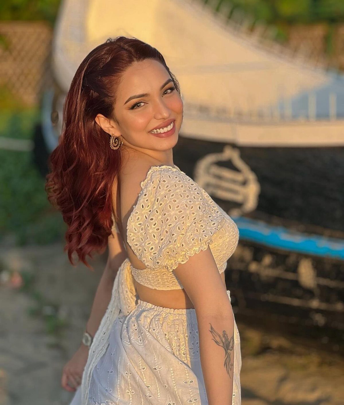 A smiling woman with reddish hair wearing a white eyelet lace crop top and skirt.