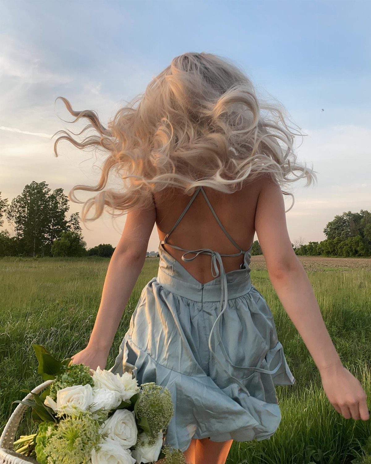 A girl with wavy blonde hair wearing a light blue satin slip dress in a grassy field.