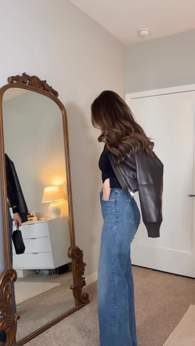 Woman wearing blue flare jeans and a black top with a leather jacket draped over her shoulders.