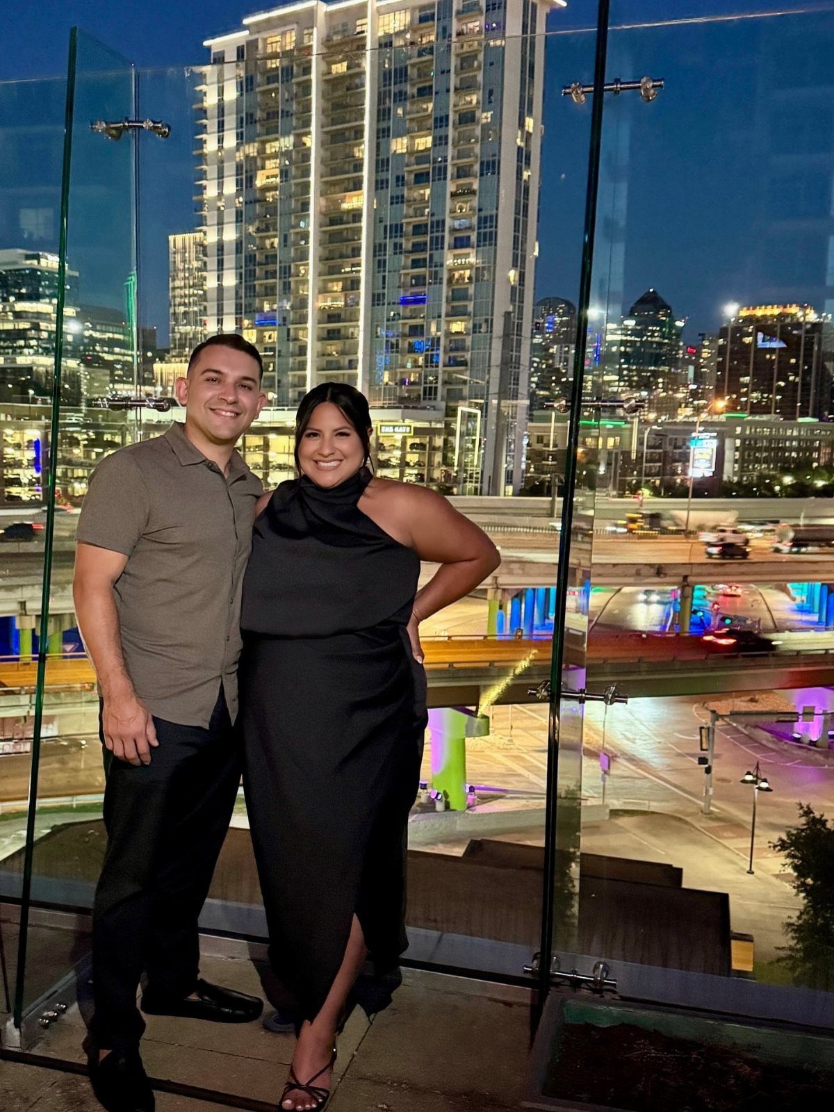 A man and a woman smiling while posing on a balcony overlooking a city skyline at night.