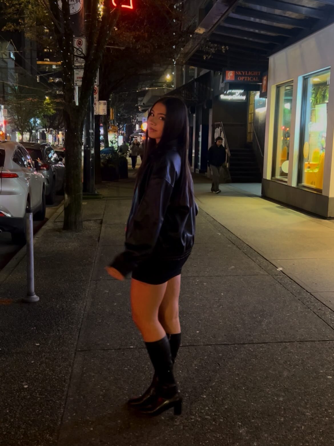 Woman in a black jacket, mini skirt, and tall boots standing on a city sidewalk at night.