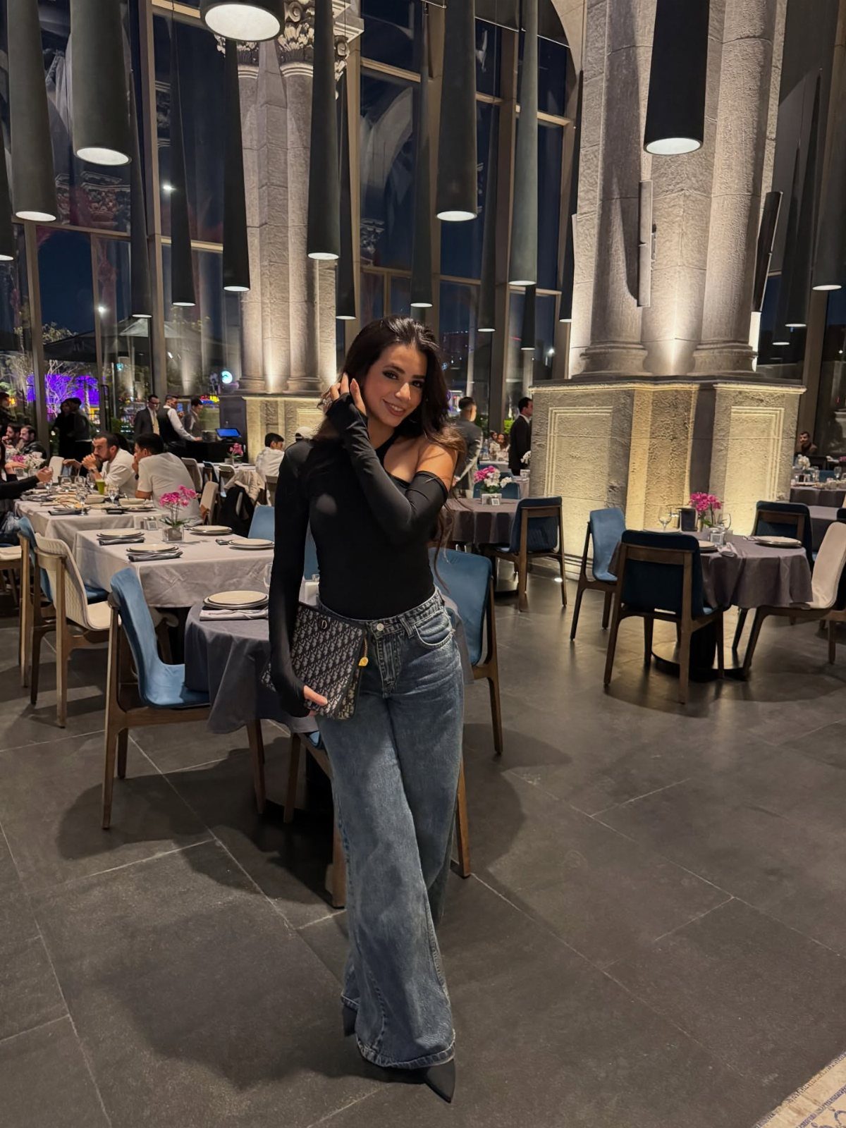 Woman posing in a black bodysuit and wide-leg blue jeans inside an upscale restaurant.