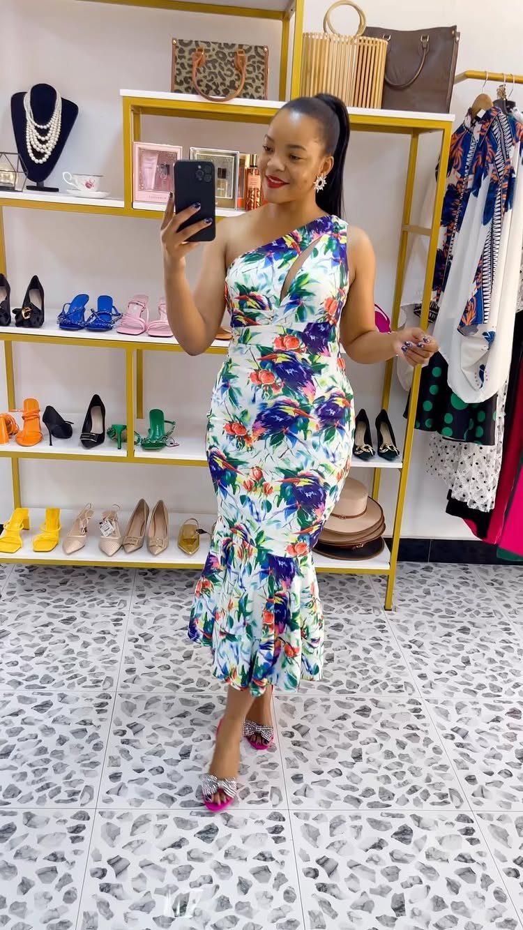 Woman posing in a colorful floral one-shoulder maxi dress in a clothing boutique.