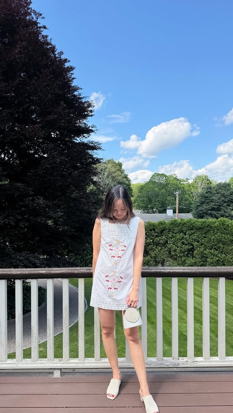 Woman in a white embroidered mini dress standing on a wooden deck outdoors.
