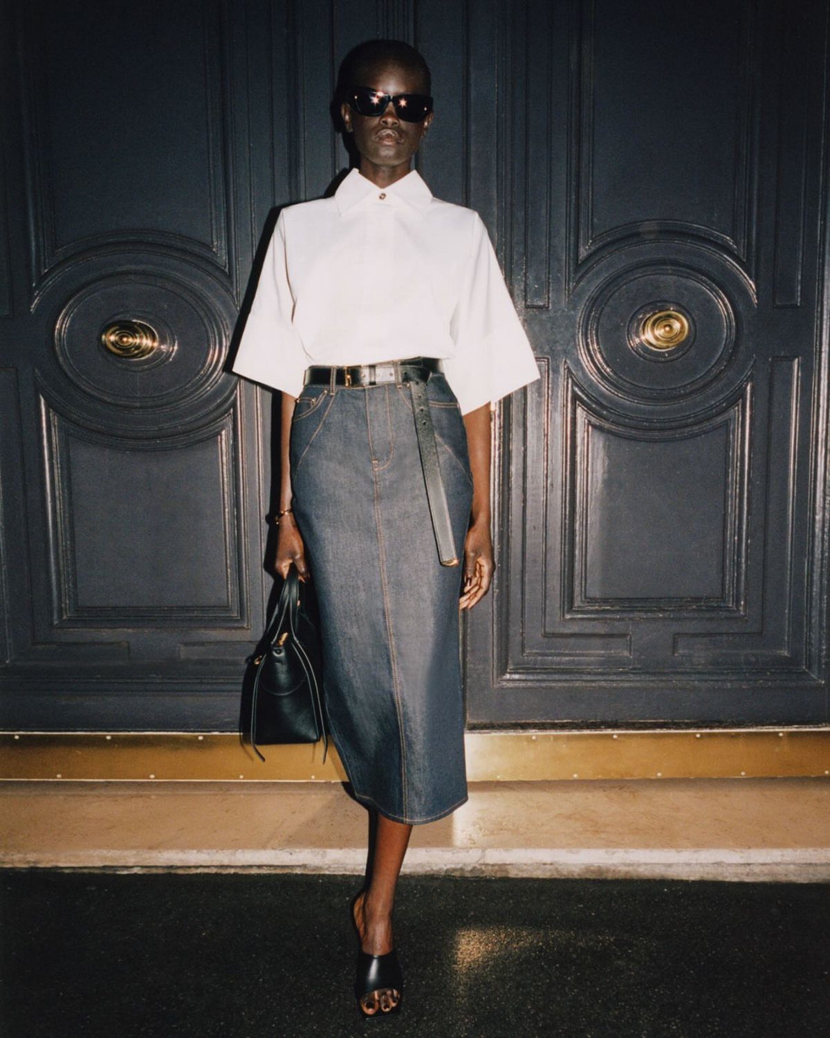 A person posing in a white blouse and a long dark denim pencil skirt.