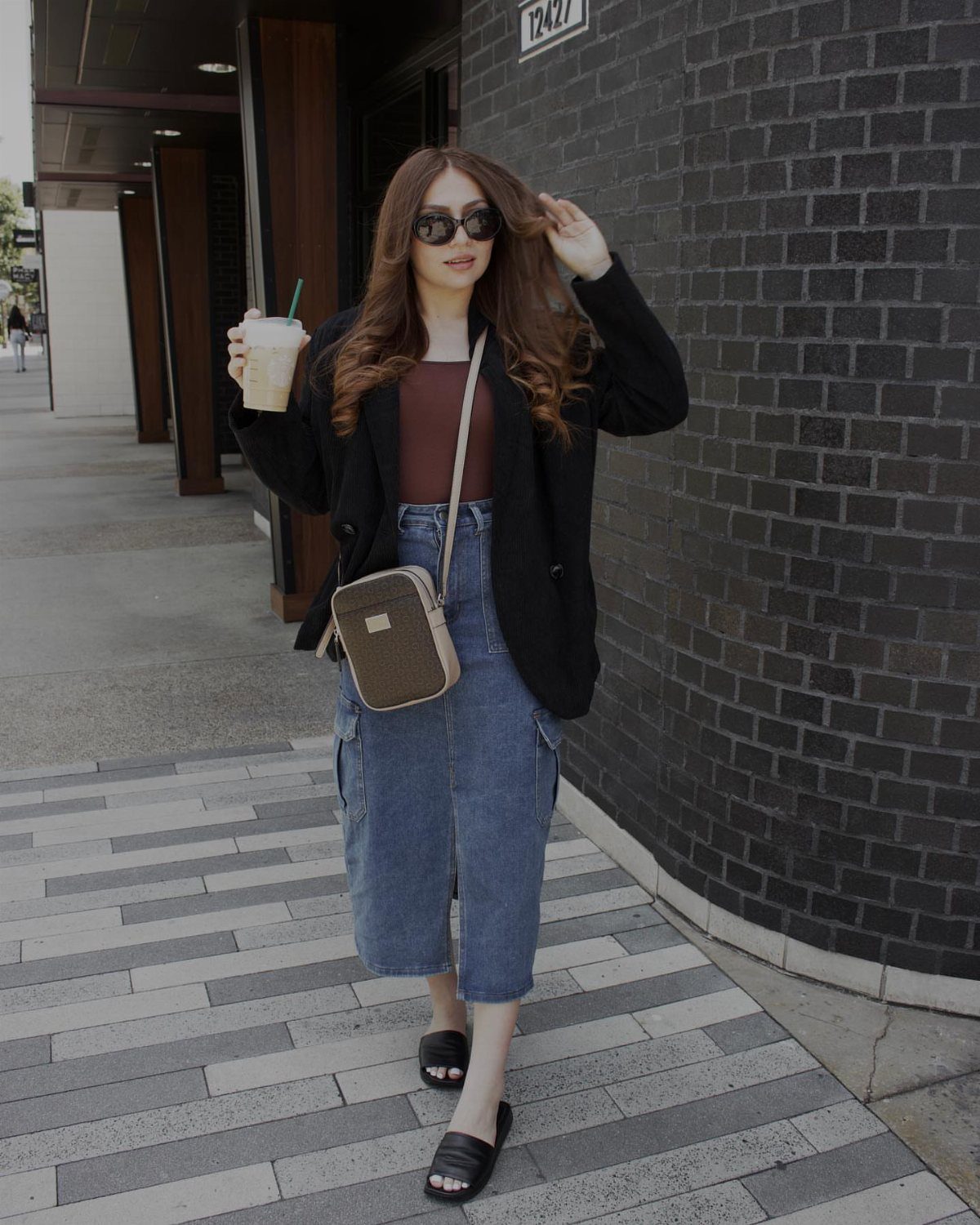 Woman walking in a black blazer, brown top, and blue denim midi skirt while holding coffee.