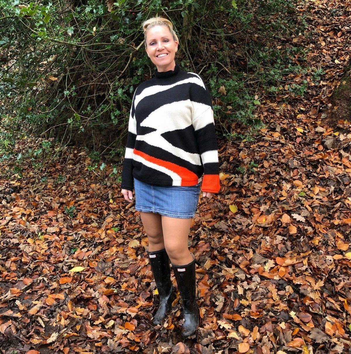 A woman in a patterned sweater and denim mini skirt standing among autumn leaves.