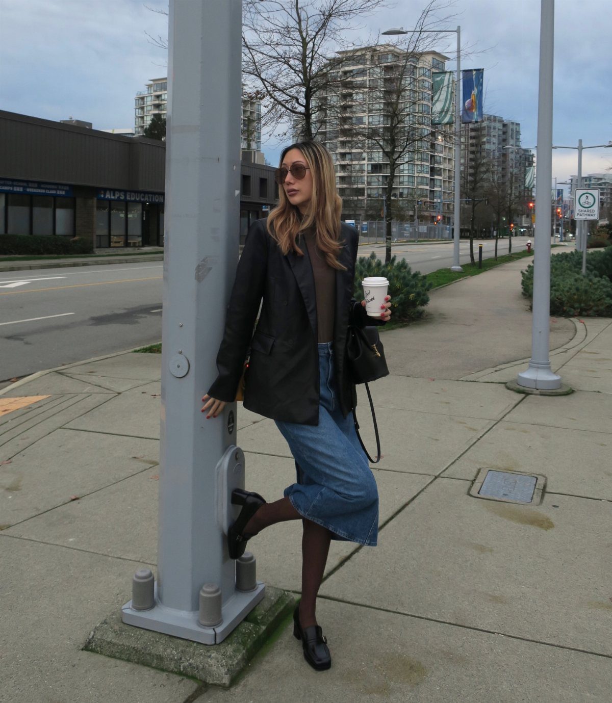 Woman wearing a black leather blazer and denim skirt standing on a city sidewalk.