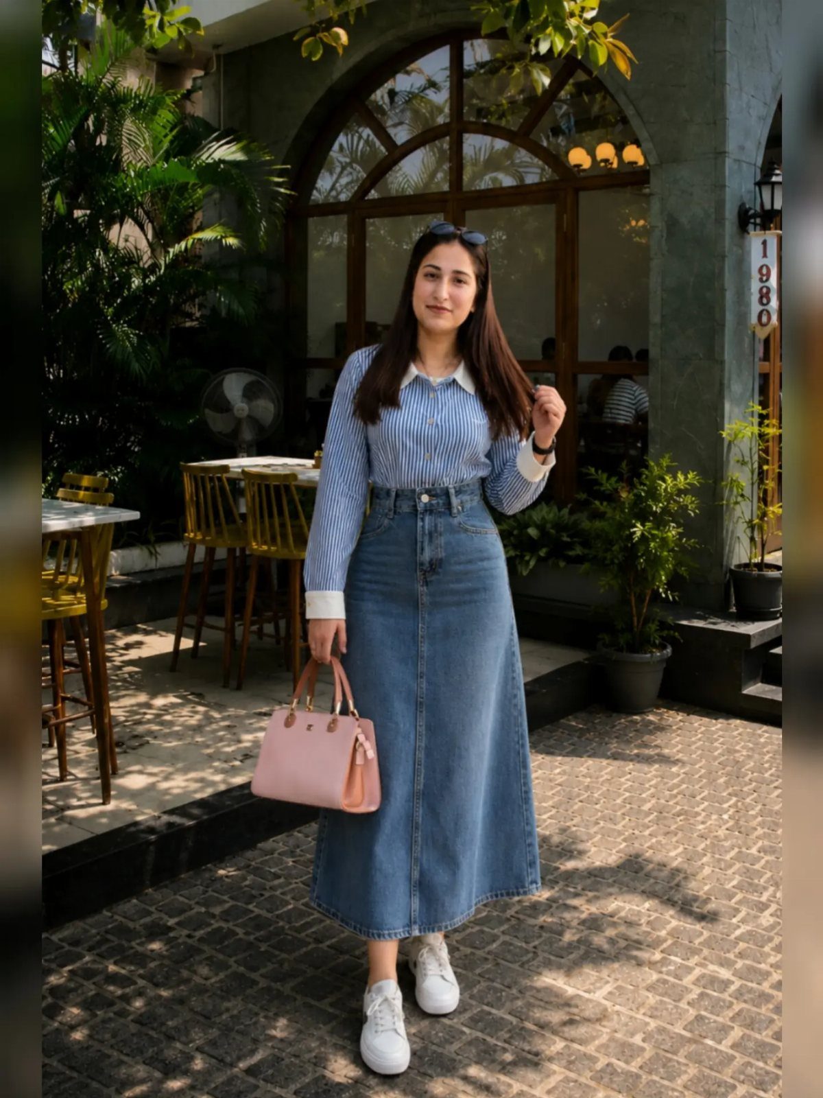 A woman wearing a long denim skirt, striped shirt, and white sneakers standing outdoors.