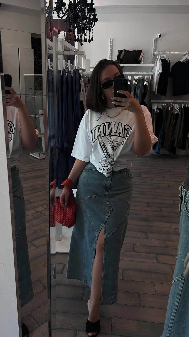Woman in a white graphic t-shirt and long denim skirt taking a mirror selfie.