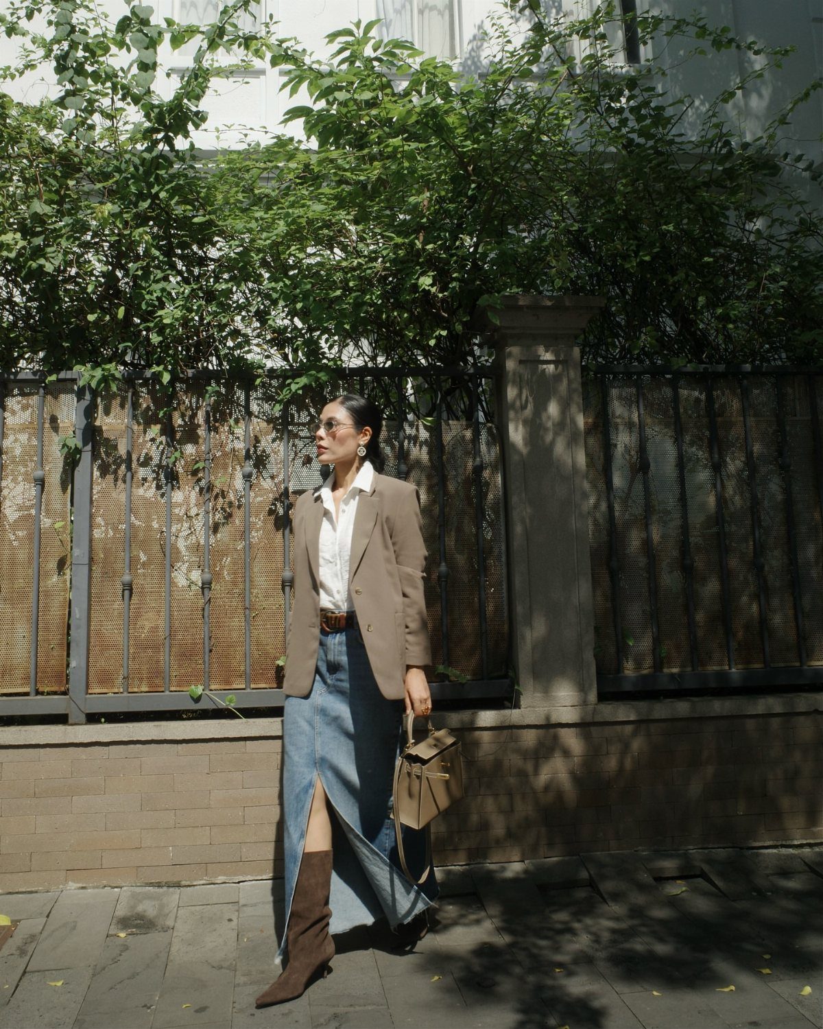 Woman wearing a long denim skirt, white shirt, and oversized tan blazer walking outdoors.