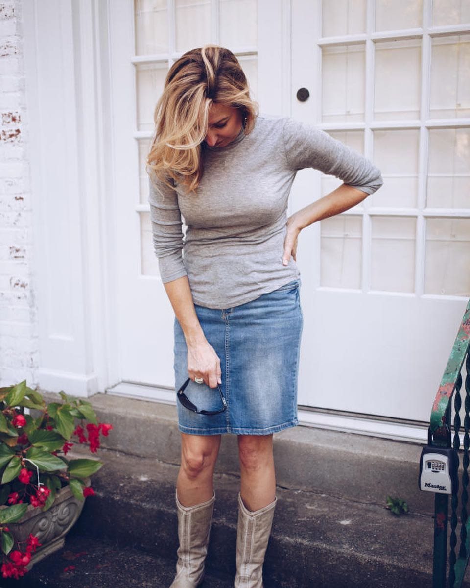 Woman wearing a grey long-sleeved shirt, blue denim skirt, and tall beige boots standing by a white door.