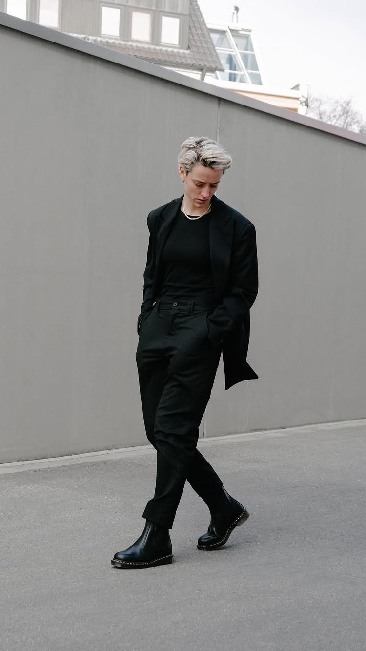 A person walking in an all-black pantsuit and black leather boots against a gray wall.