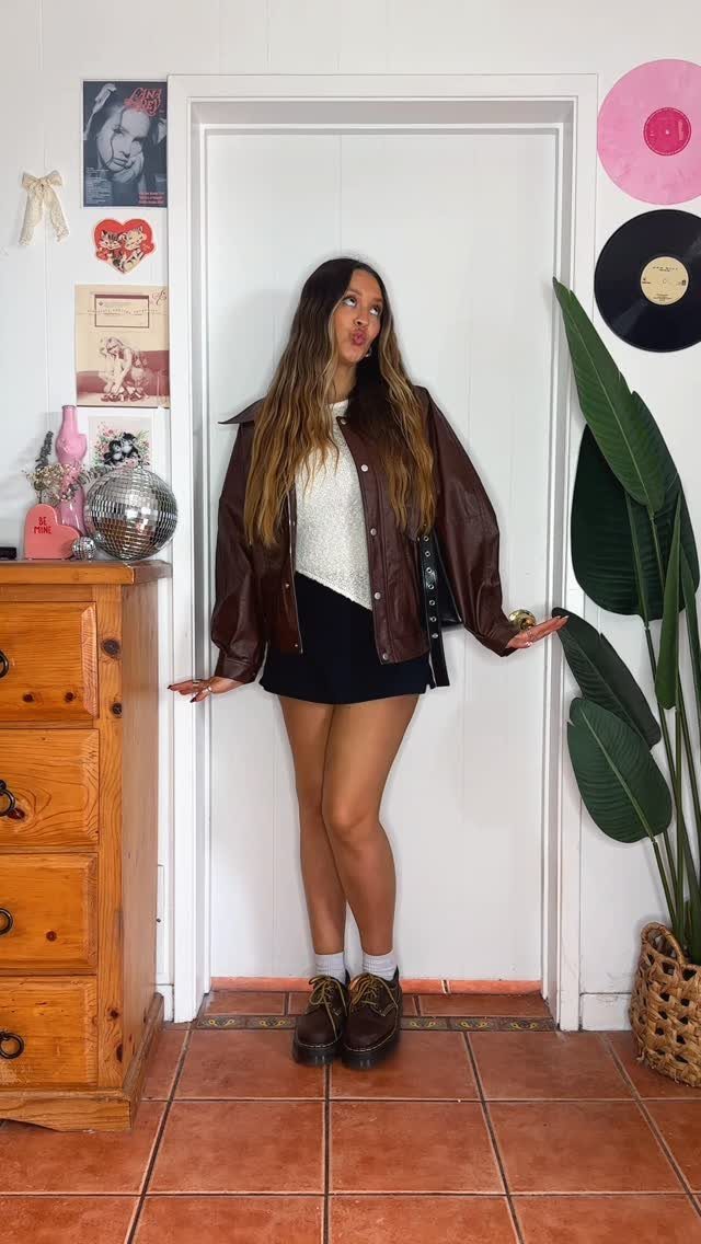 A woman posing in a leather jacket, black skirt, and Doc Marten boots in a room.