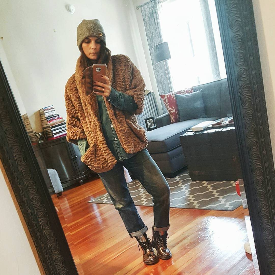 Woman in a brown furry coat, denim shirt, jeans, and boots taking a mirror selfie.