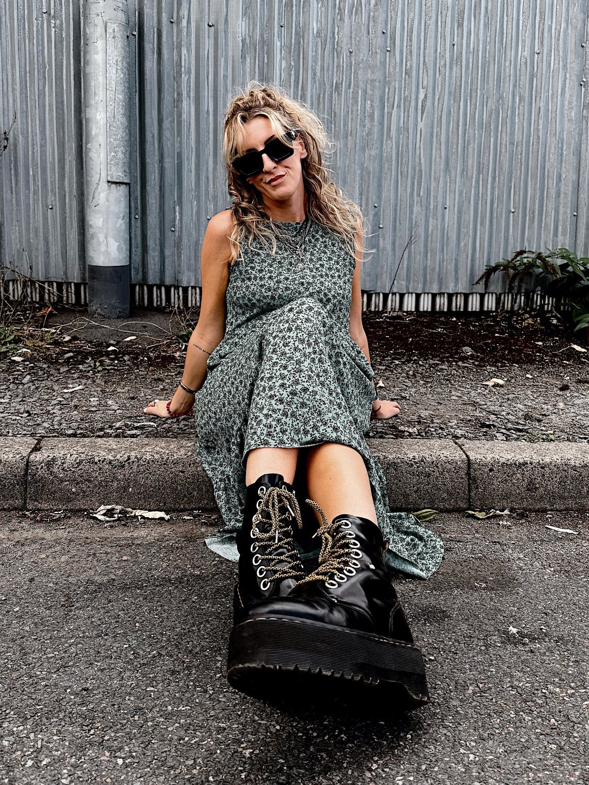 Woman in a floral dress and black platform boots sitting on a curb outdoors.