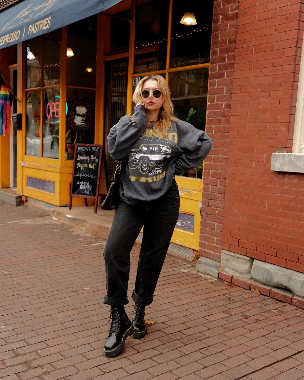Woman in a graphic sweatshirt, black baggy jeans, and combat boots standing on a brick sidewalk.