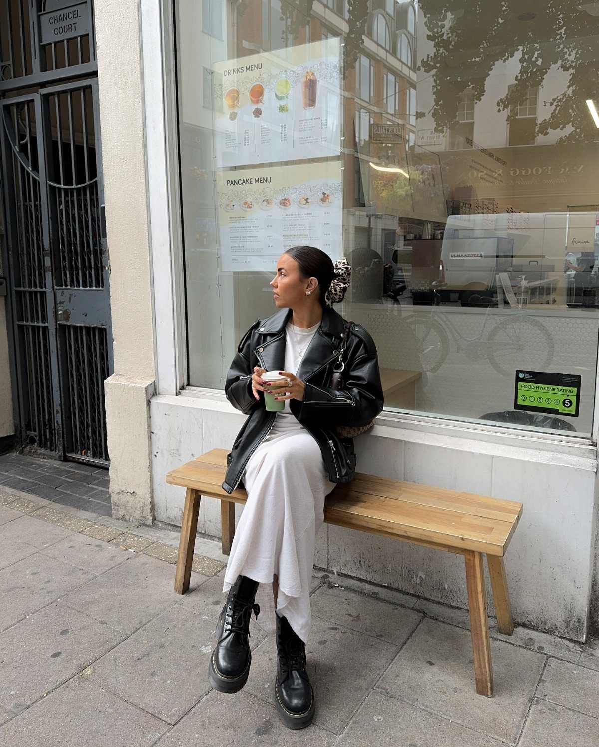 Woman in a black leather jacket, white skirt, and black boots sitting on a wooden bench.