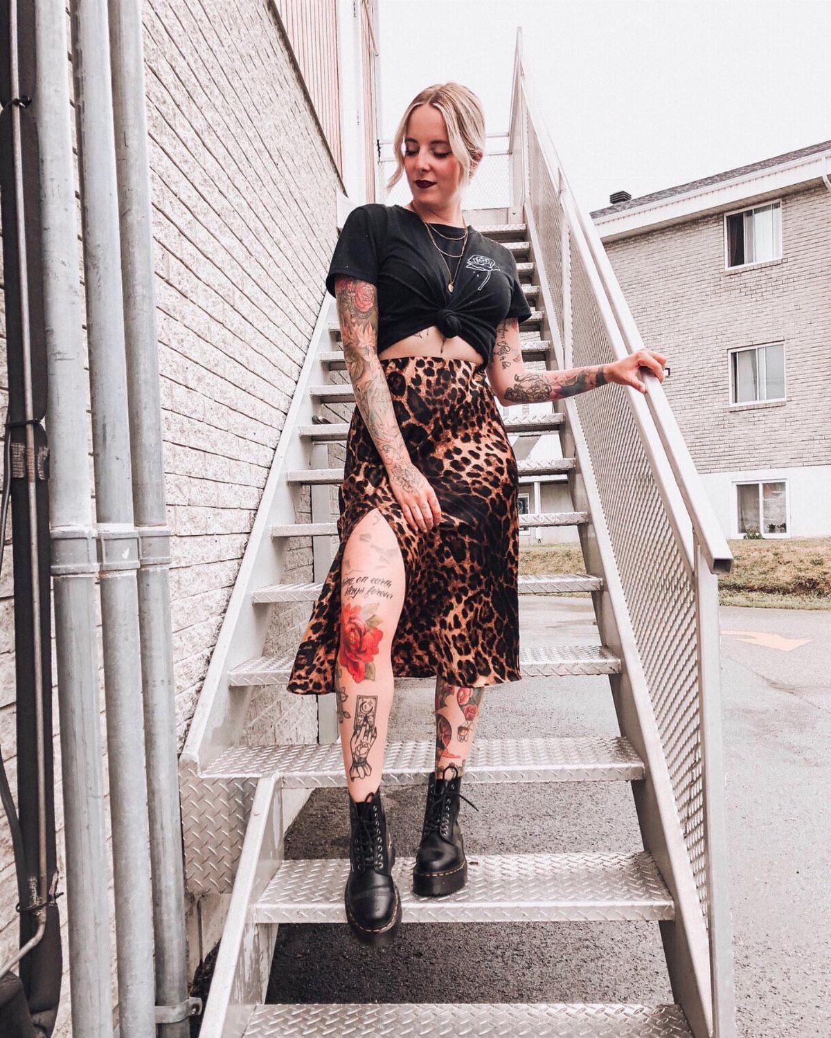 A tattooed woman wearing a leopard print skirt and black combat boots standing on metal stairs.