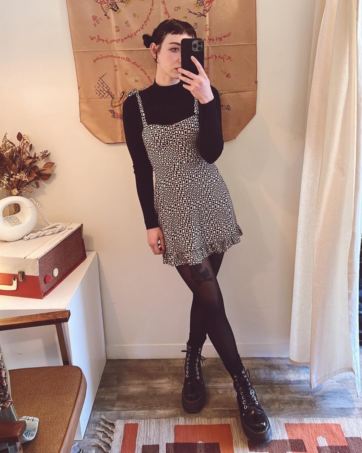 Woman in a patterned slip dress, black turtleneck, tights, and combat boots taking a selfie.