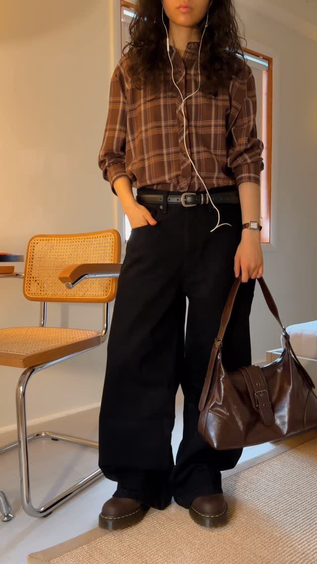 Woman wearing a brown plaid shirt, black wide-leg trousers, and dark brown Doc Marten boots.