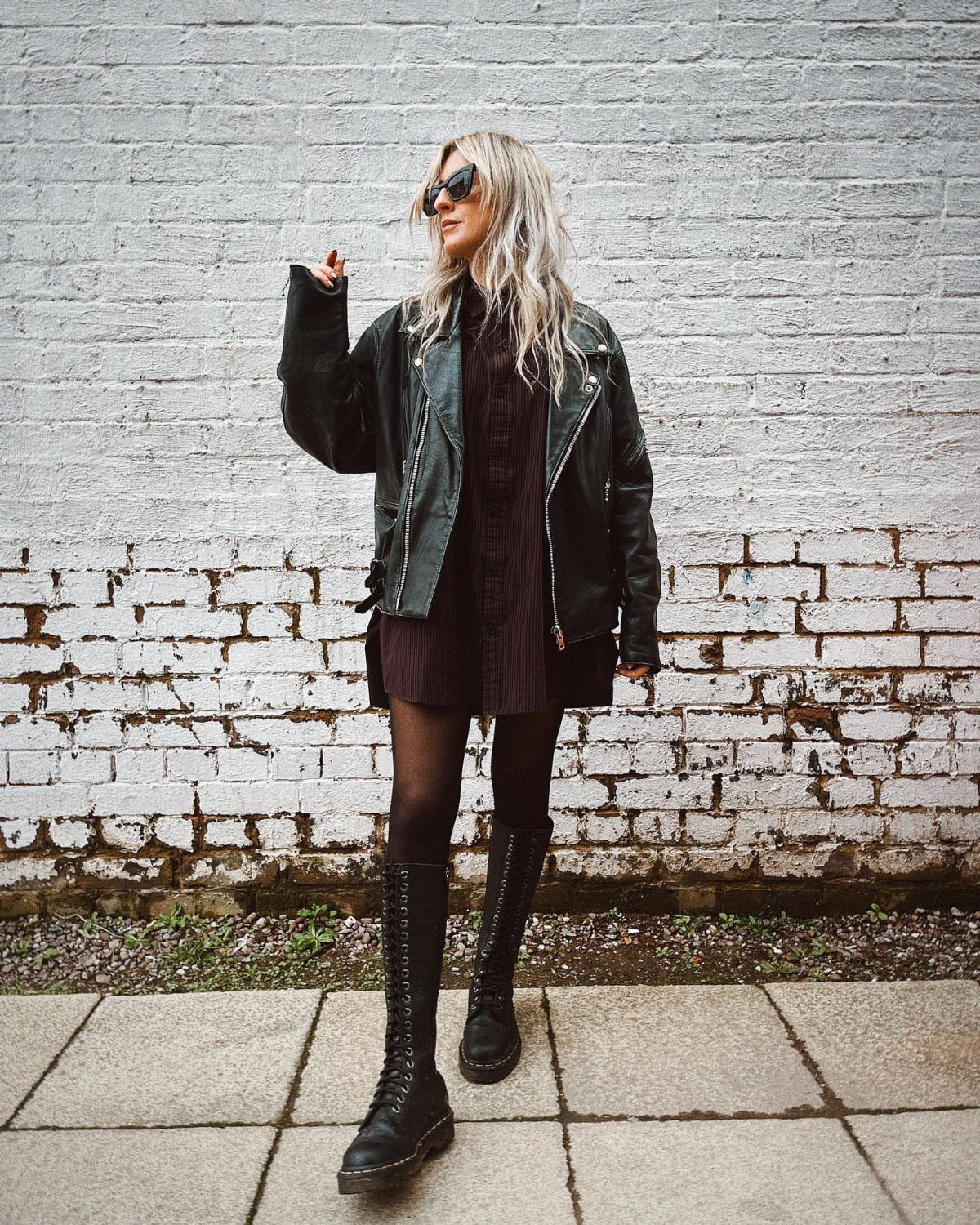 Blonde woman in a leather jacket, ribbed knit minidress, and tall black boots posing against a brick wall.