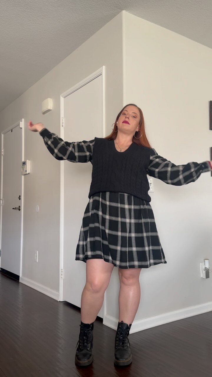 Woman posing in a black sweater vest, plaid dress, and black combat boots.