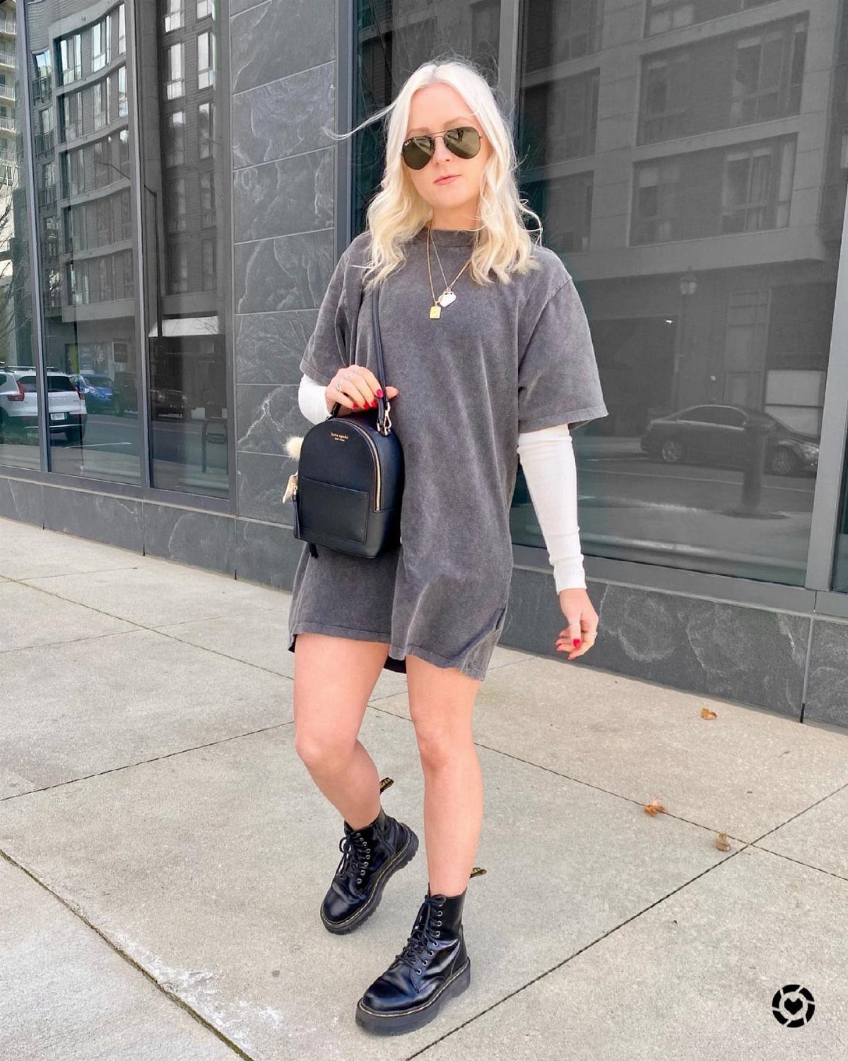 Blonde woman in a grey oversized t-shirt dress and black boots standing on a city sidewalk.