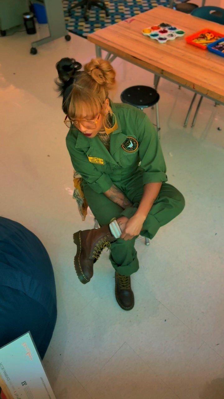 Person in a green utility jumpsuit sitting and adjusting dark brown lace-up boots.