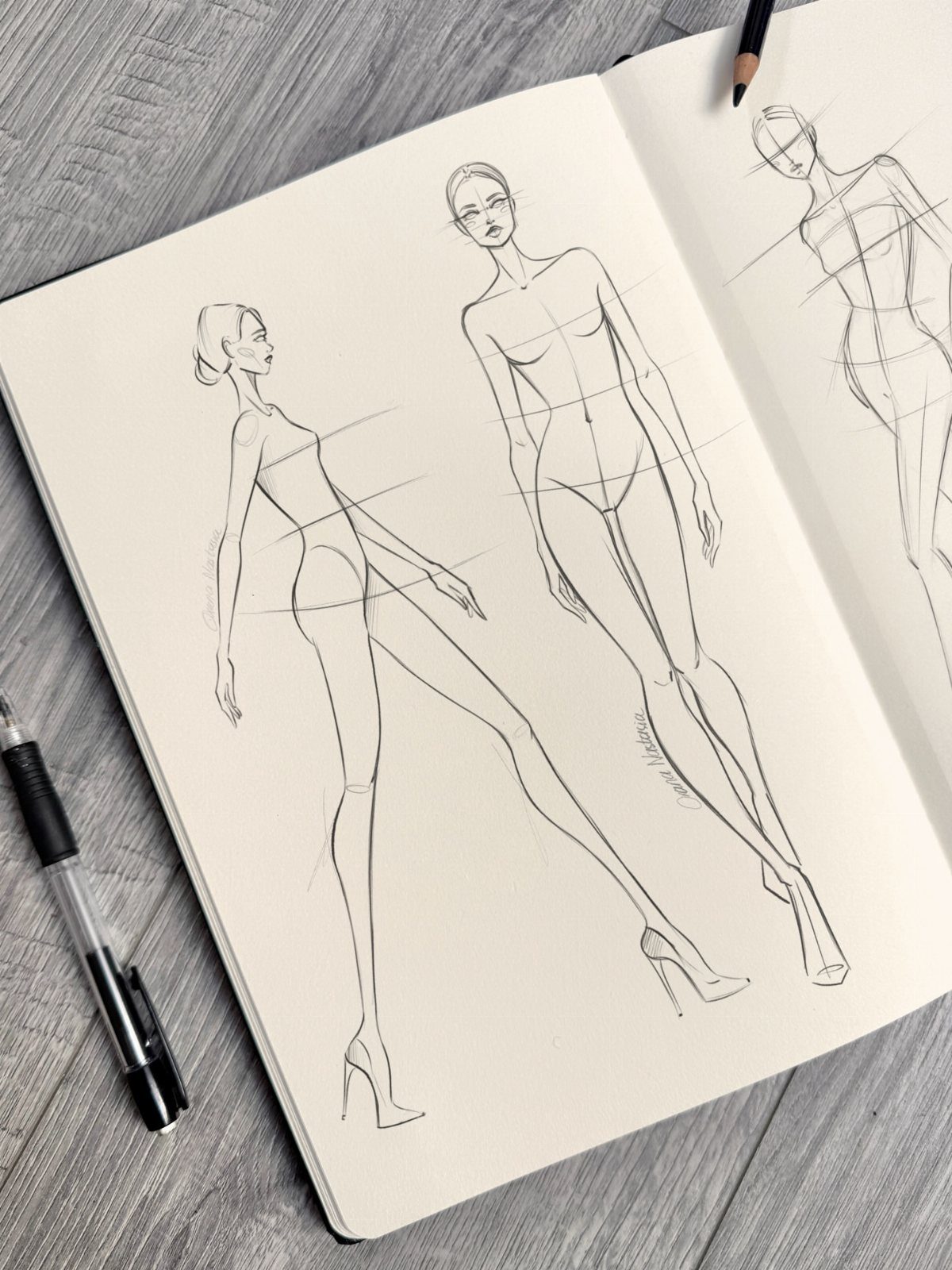 Pencil sketches of elongated fashion croquis figures drawn in an open sketchbook on a wooden surface.