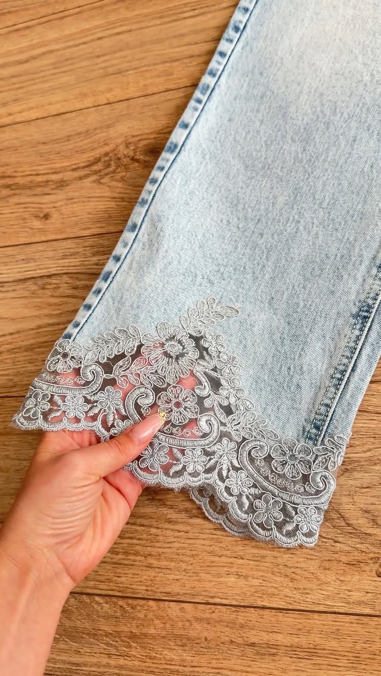 Light blue denim jeans featuring intricate grey lace embroidery along the bottom hemline.