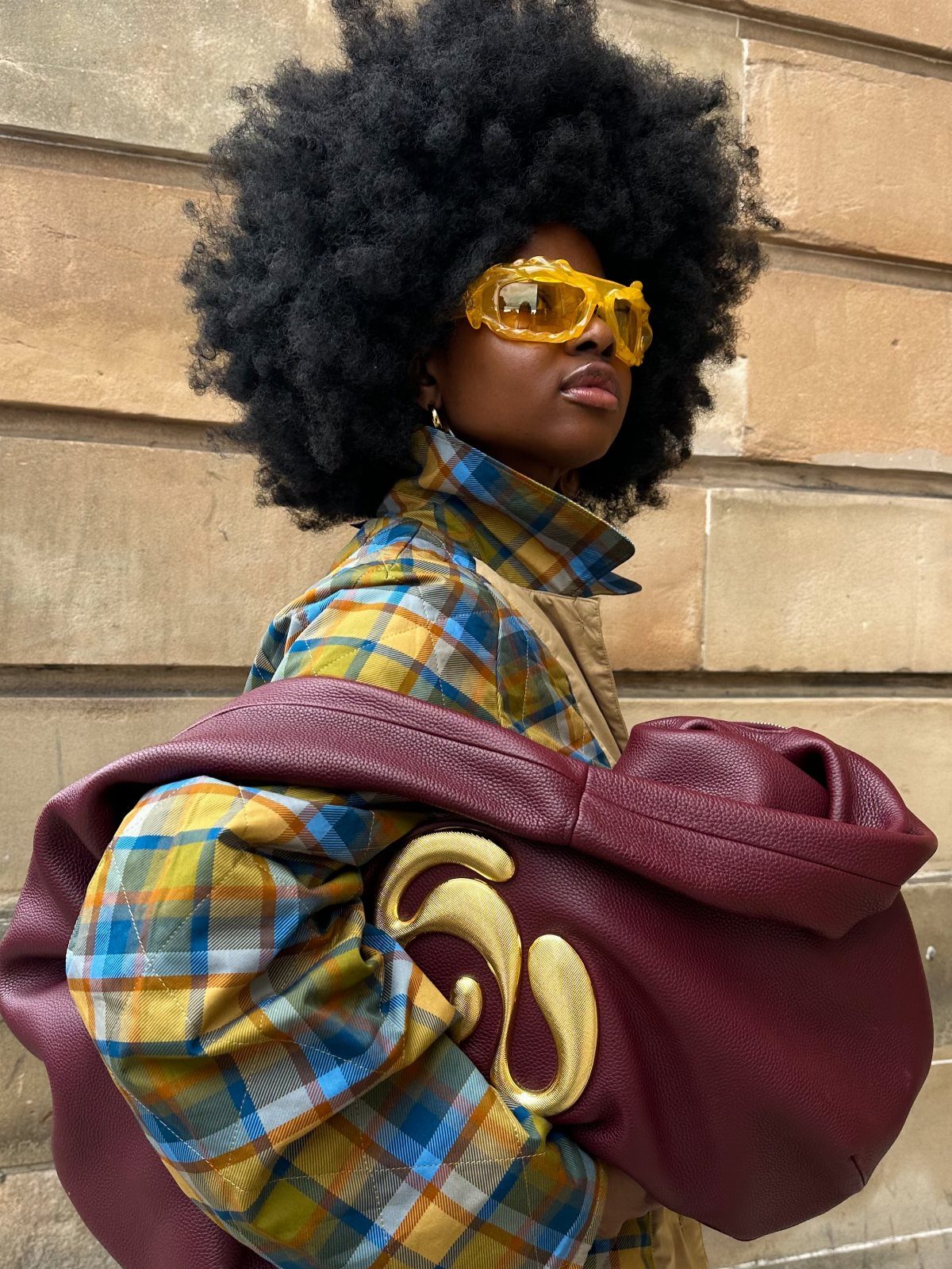 Person with large afro wearing yellow sunglasses and plaid shirt carrying a burgundy leather bag.