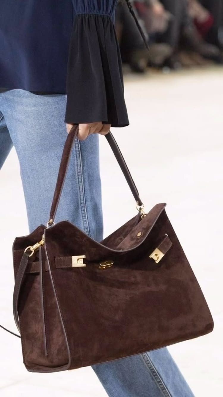 A large dark brown suede tote bag with gold hardware held by a person.