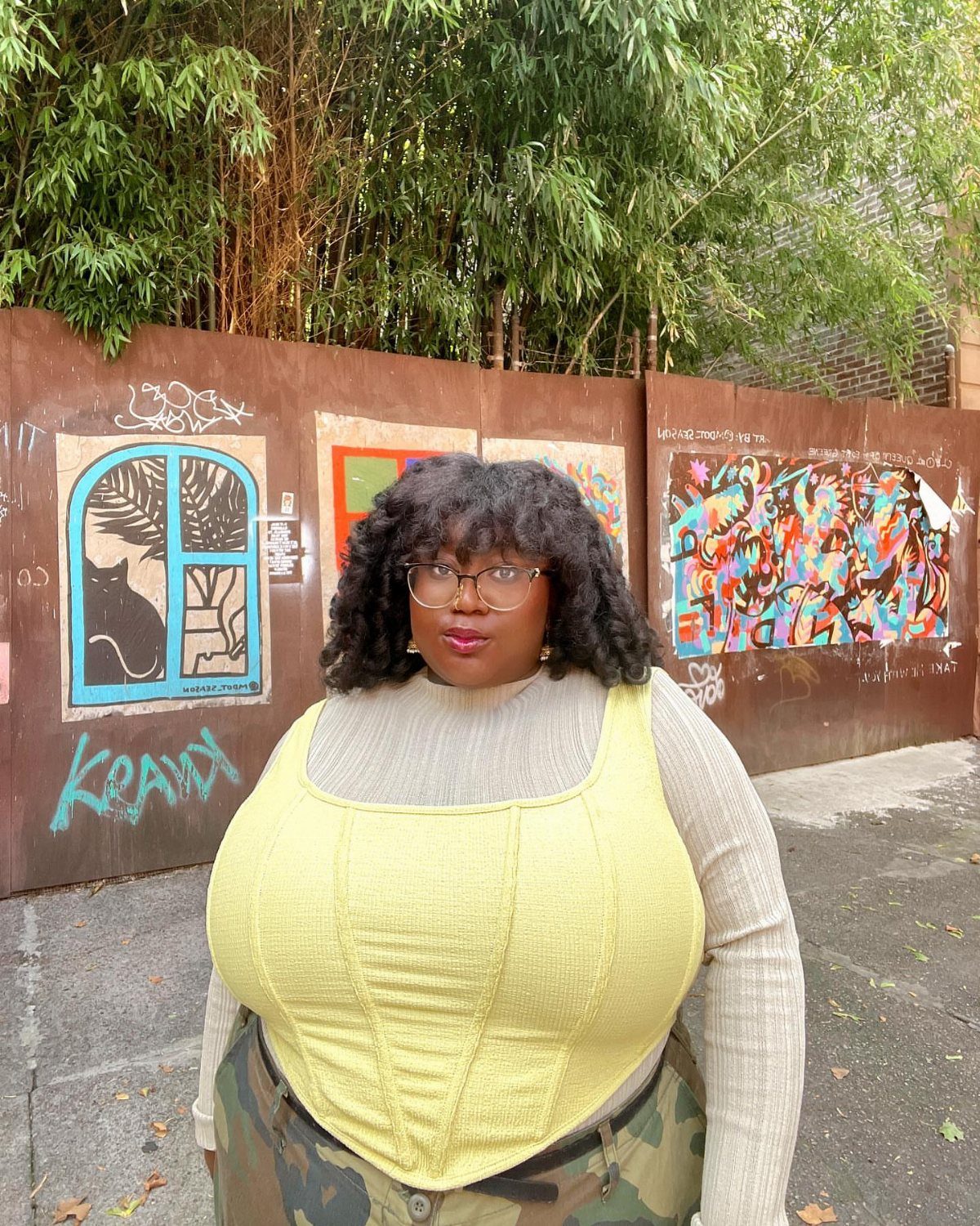 Woman wearing a yellow corset top, white turtleneck, and camouflage cargo pants against a graffiti wall.