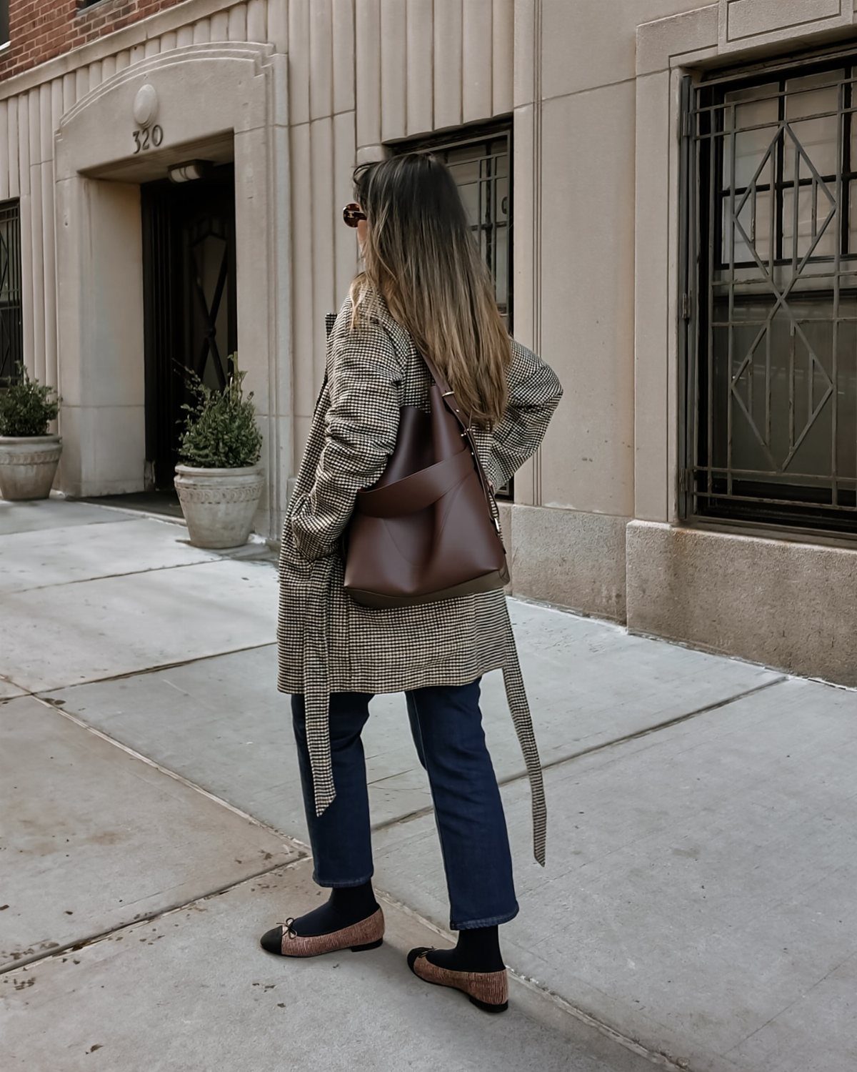 Woman wearing a plaid coat, blue jeans, and ballet flats walking on a city sidewalk.